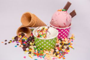 "Two scoops of ice cream in colorful polka-dot cups, topped with sprinkles, surrounded by chocolate chips, candy pieces, and empty waffle cones."
